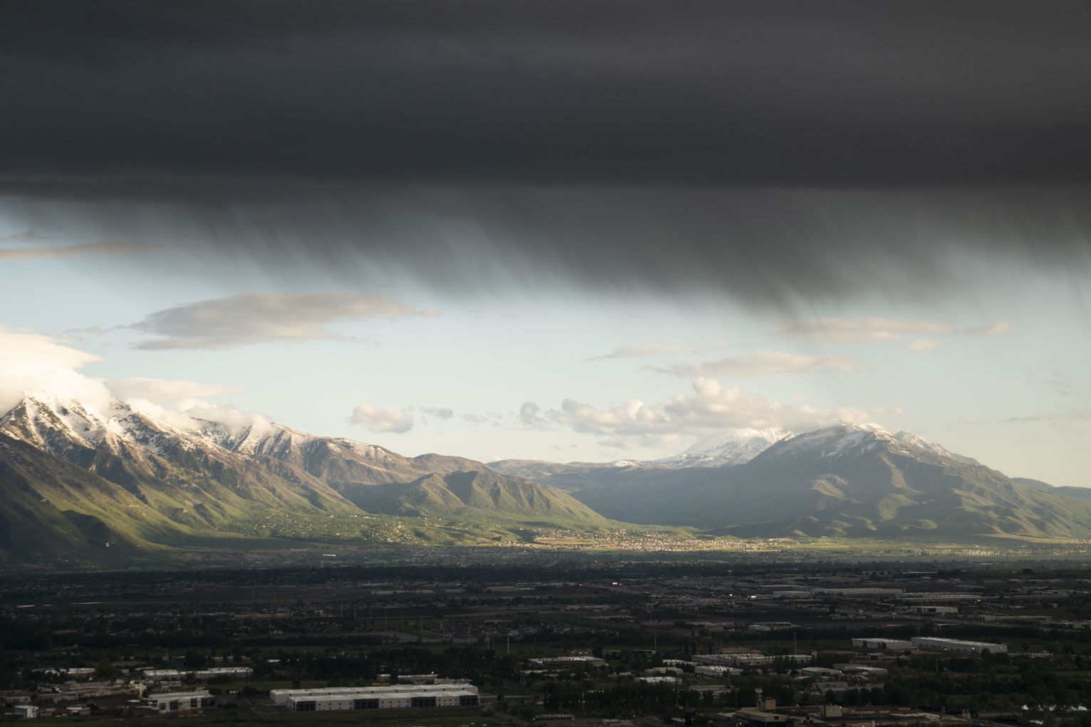 A great flat heavy dark cloud rains a bit on a town and mountainside that are catching sunlight somehow